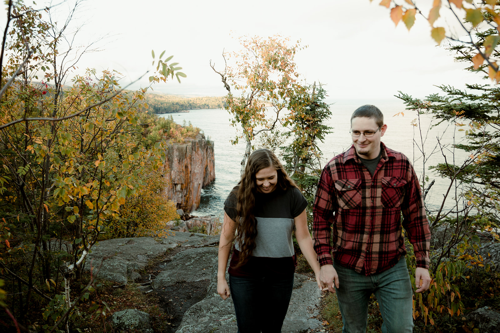 Man and woman walking hand in hand on rocky terrain with autumn leaves and lake in the background.